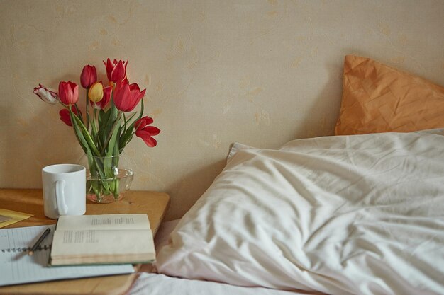 Wooden table with paper notebook book cup of tea and spring flowers next to bed