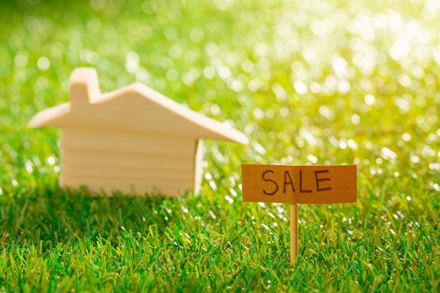 Wooden toy house on grass with sale sign close up