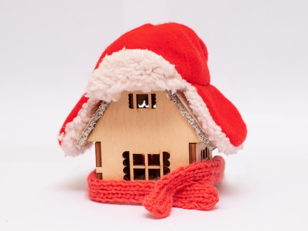 A wooden toy house in the snow in winter is covered with a hat and wrapped in a scarf The concept of keeping warm