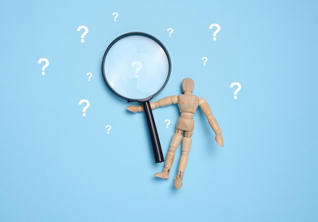 a wooden toy puppet holds a magnifying glass and question marks on a blue background concept of answering questions and gaining knowledge