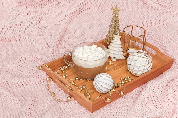 Wooden tray with cup of cocoa, marshmallows and christmas decorations on warm knitted pink plaid. 