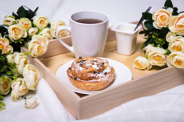 Wooden tray with sweet bun, tea, yoghurt and flowers