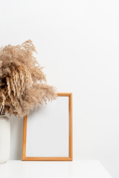 Wooden vertical frame with white vase of dry flowers over white wall