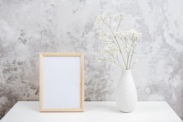 Cornice vuota bianca verticale in legno e bouquet di gypsophila bianchi piccoli fiori in vaso sul tavolo sul muro di cemento grigio