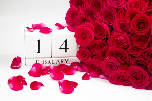 Wooden white calendar February 14 with a bouquet of red roses and petals on a white background.