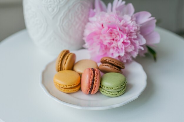 Woonkamer interieur. Boeket van roze pioenrozen in een vaas, veelkleurige Franse macaron gebakjes. Knus. Voorjaar.