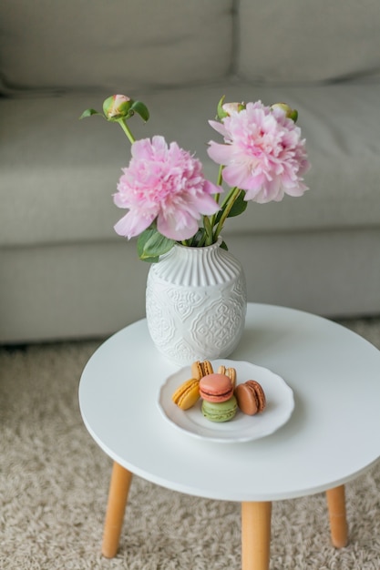 Woonkamer interieur. Boeket van roze pioenrozen in een vaas, veelkleurige Franse macaron gebakjes. Knus. Voorjaar.