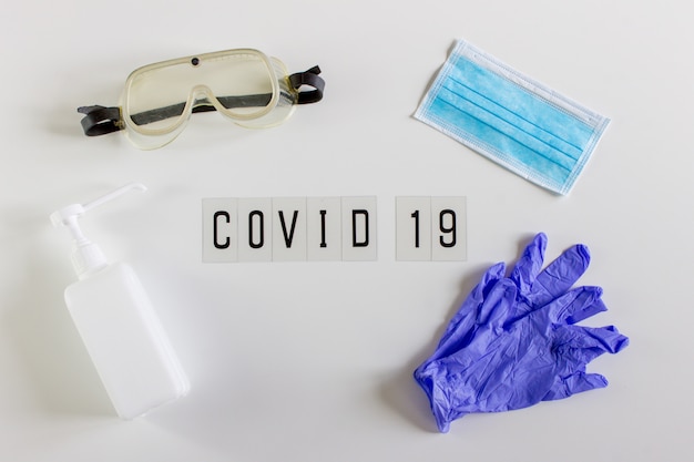 Woord "Covid 19" liggend op een witte achtergrond. Omringd door een veiligheidsbril, alcoholgel, handschoenen en masker.