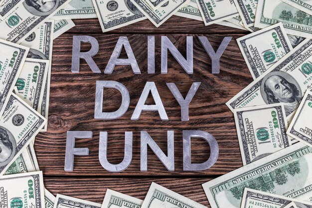 Woorden RAINY DAY FUND gelegd op een houten oppervlak met metalen letters omringd door Amerikaanse dollarbankbiljetten