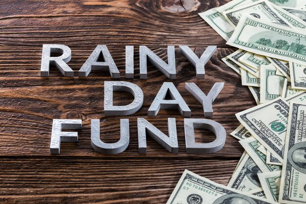 Woorden RAINY DAY FUND gelegd op houten oppervlak met metalen letters met regendruppels en Amerikaanse dollar bankbiljetten recht perspectief