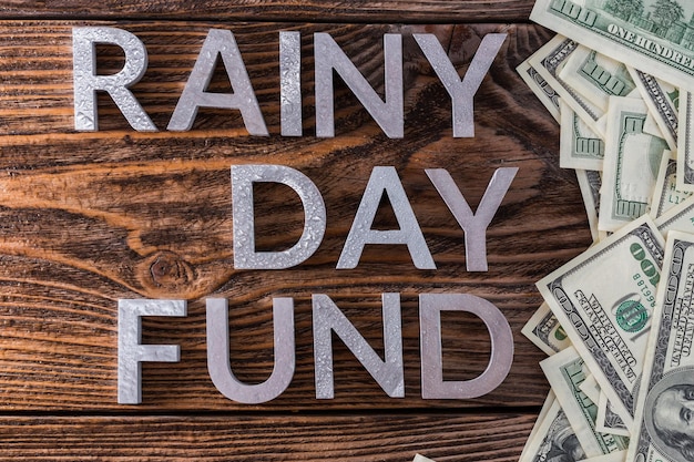 Woorden RAINY DAY FUND gelegd op houten oppervlak met metalen letters met regendruppels en Amerikaanse dollarbankbiljetten