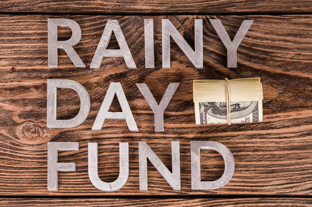 Woorden RAINY DAY FUND op houten oppervlak gelegd door metalen letters met een kleine rol Amerikaanse dollarbankbiljetten plat boven het zicht