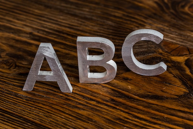 흰색 금속 문자가 있는 ABC라는 단어는 선택적 초점이 있는 짙은 갈색 나무 배경에 서 있습니다.