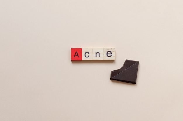 the word "Acne" is written with wooden tiles with a red "A" with a bitten piece of chocolate on a beige background