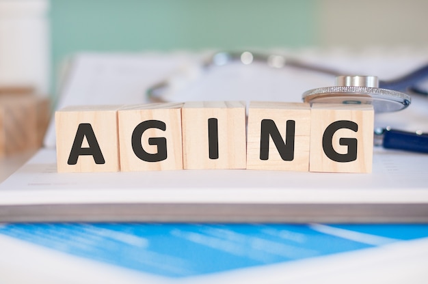 The word AGING is written on wooden cubes near a stethoscope on paper . Medical concept.