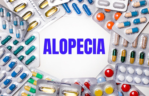 The word ALOPECIA is written on a light background surrounded by multicolored packages with pills Medical concept