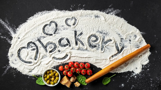 Фото Слово пекарня и сердце написано на муке со скалкой и ингредиентами для приготовления итальянской еды, вид сверху. абстрактный фон выпечки