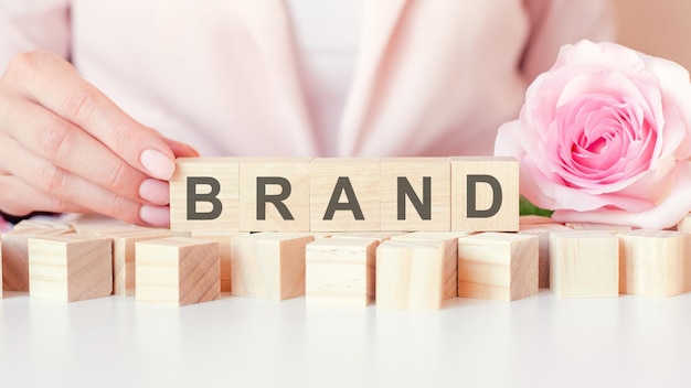 The word brand is made up of letters on wooden blocks. a woman's hand holds a cube on a pink background. flower rose in the background