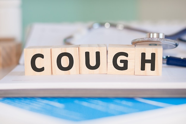 The word cough is written on wooden cubes near a stethoscope on a paper background. Medical concept.