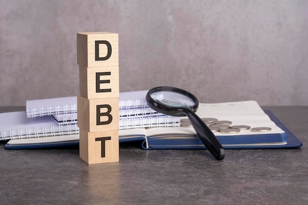 The word Debt is written on wooden cubes on a gray background. close-up of wooden elements, magnifying glass, paper documents and coins on a notebook