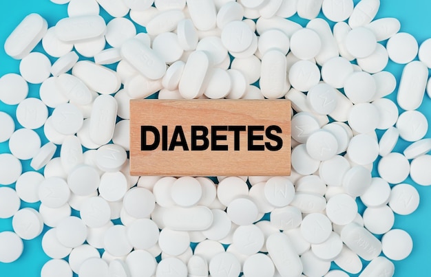 The word Diabetes is written on wooden blocks on pile of white pills on blue background.