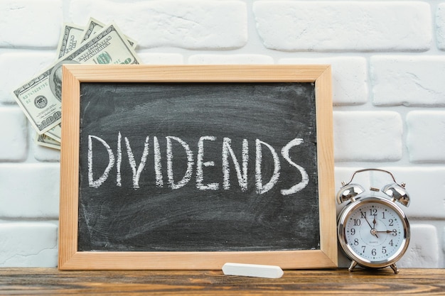 Photo the word dividends on a black chalkboard in a frame standing on a white brick wall dollar bills and an alarm clock nearby