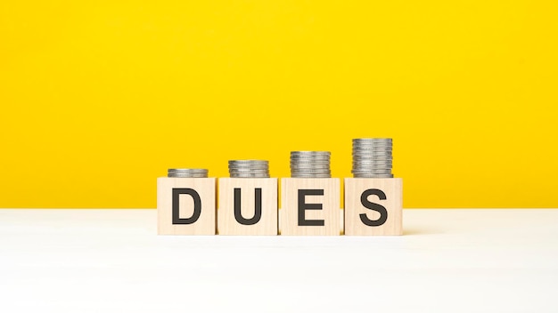 Word dues on wooden blocks in yellow background with increasing stack of coins