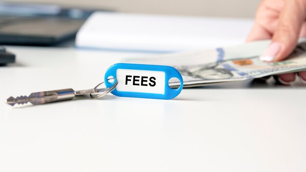 The word fees is written on a blue keychain. The key is on the office desk. in the background, a woman's hand is holding money bills
