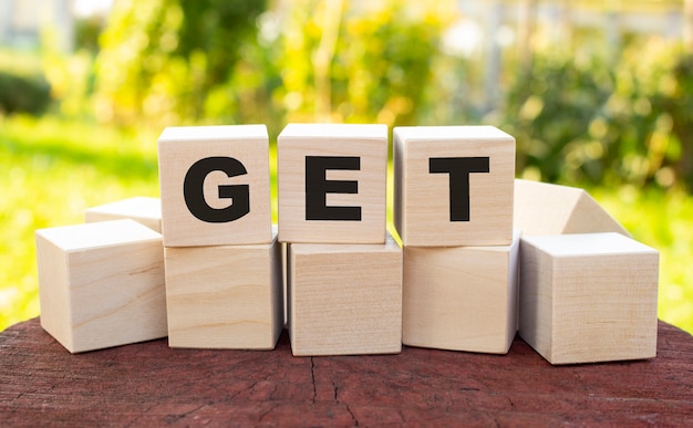 The word GET is made up of wooden cubes lying on an old tree stump against a blurred garden background.