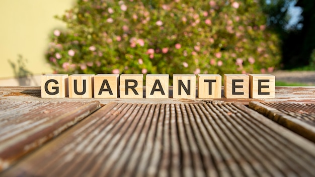 The word guarantee is written on wooden cubes. the blocks are placed on an old wooden board illuminated by the sun. in the background is a brightly blooming shrub