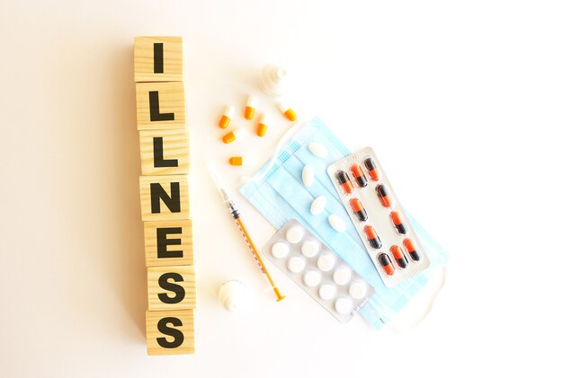 The word ILLNESS is made of wooden cubes on a white background with medical drugs and medical mask.