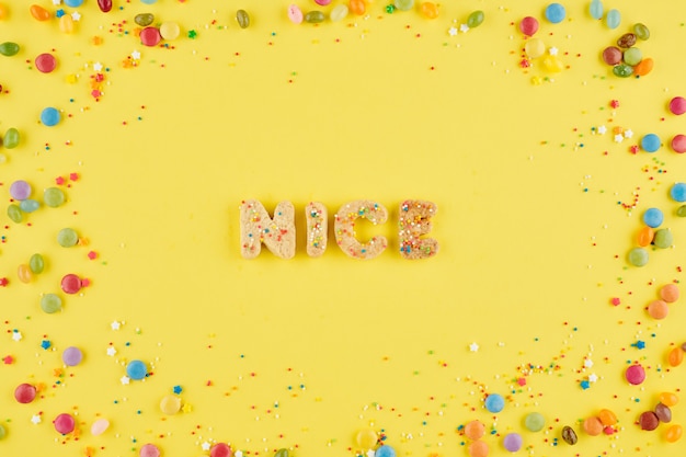 Word nice created with homemade sugar cookies on yellow background decorated with candies and sprinkles