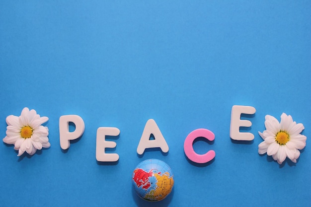 Photo word peace made by letters near little figure of a globe and a flowers of white chrysanthemums blue background and empty space to insert text