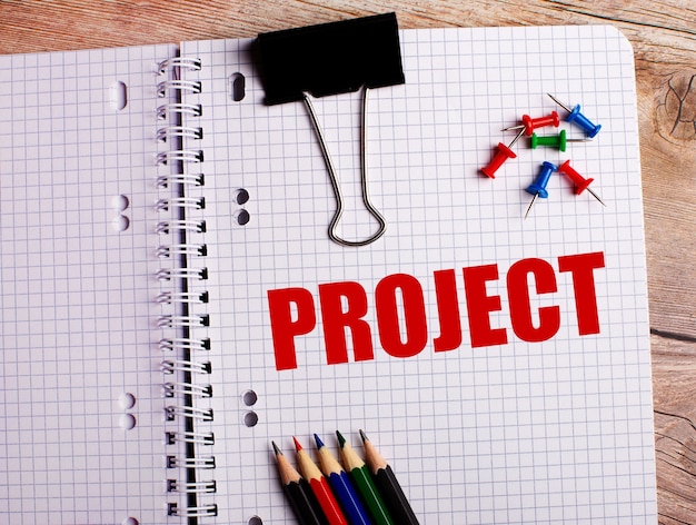 The word PROJECT is written in a notebook near multi-colored pencils and buttons on a wooden table.