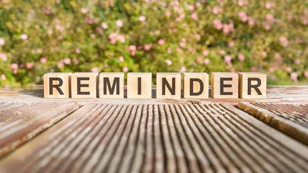 The word REMINDER is written on wooden cubes. the blocks are placed on an old wooden board illuminated by the sun. in the background is a brightly blooming shrub