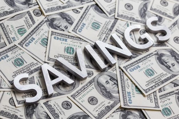 Слово SAVINGS, выложенное белыми металлическими буквами на фоне американских долларовых банкнот с избирательным фокусом