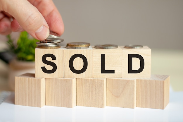 The word sold written on wood cubes. a man's hand places the coins on the surface of the cube. green potted plant on the background. Business and finance concept.