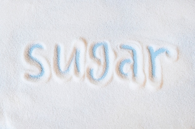 Слово сахар рука надписи на россыпи рафинированного белого сахара. Гранулы кристаллов свекольного сахара на синем фоне