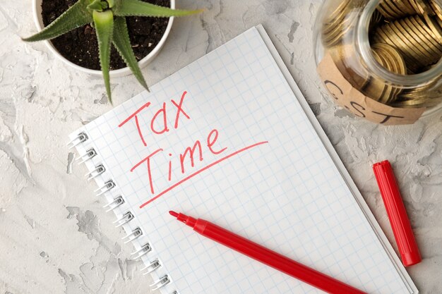 Слово налоговое время в записной книжке с банкой монет и цветком на светлом фоне бетона. вид сверху