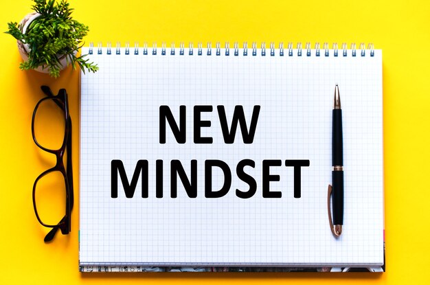Текст слова NEW MINDSET на белой карточке, черными буквами. Ручка, очки и зеленый цветок на желтой стене. бизнес-концепция. Концепция образования.