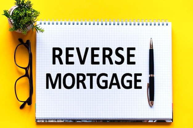 백서 카드, 검은 글자에 Word 텍스트 REVERSE MORTGAGE. 노란색 벽에 펜, 안경 및 녹색 꽃. 비즈니스 개념. 교육 개념.