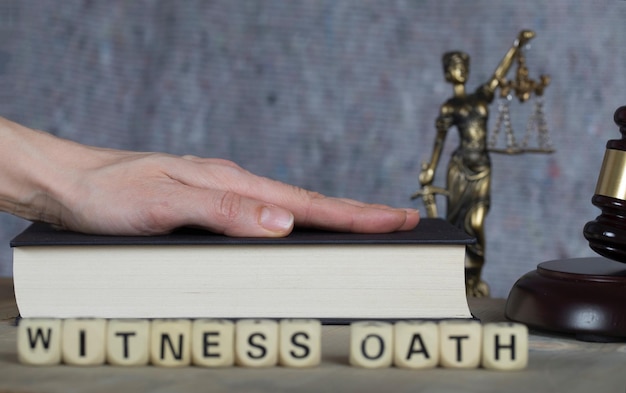 写真 木製の文字で構成された witness oath という単語。テミス像、裁判官の小槌、バック グラウンドで聖書