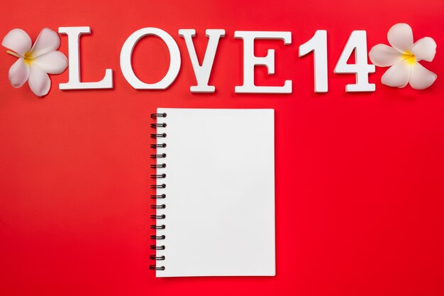 The words "14" and "love" carved from wood, plumeria flowers, red background, a clean open notebook. Valentine's day.