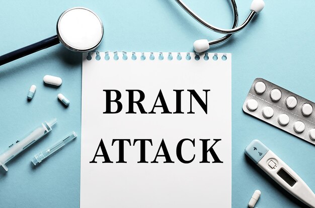 The words BRAIN ATTACK written on a white notepad on a blue surface near a stethoscope, syringe, electronic thermometer and pills. Medical concept.