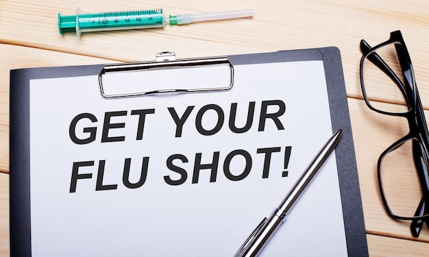 The words GET YOUR FLU SHOT is written on a white piece of paper next to black-rimmed glasses, a pen and a syringe