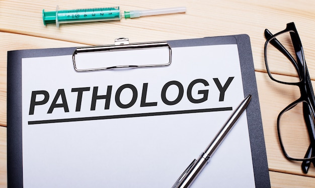 The words PATHOLOGY is written on a white piece of paper next to black-rimmed glasses, a pen and a syringe.