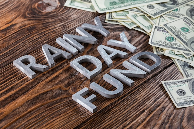 Слова RAINY DAY FUND выложены на деревянной поверхности металлическими буквами с каплями дождя и банкнотами в долларах США