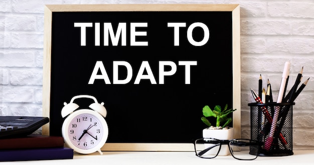 The words TIME TO ADAPT is written on the chalkboard next to the white alarm clock, glasses, potted plant, and pencils in a stand.