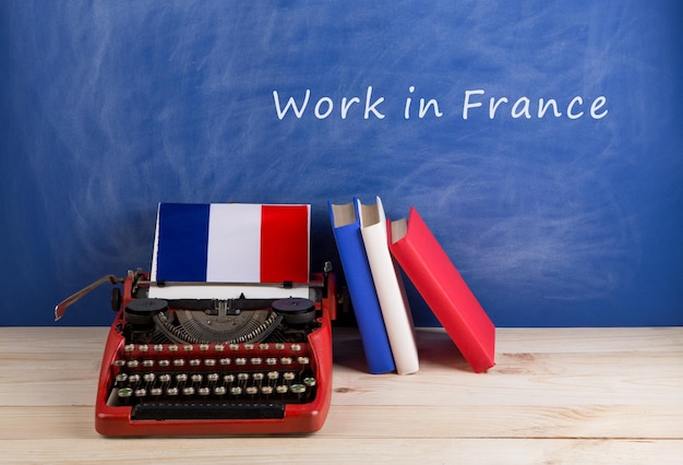 해외 작업 개념 - 빨간색 타자기, 프랑스 국기, 탁자 위의 책, "프랑스에서 일하기"라는 텍스트가 있는 칠판