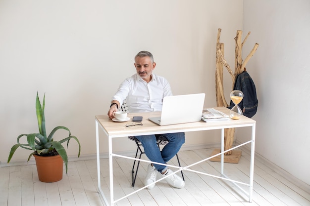 Work, break. Gray-haired stylish man sitting at table with laptop touching cup in modern bright office